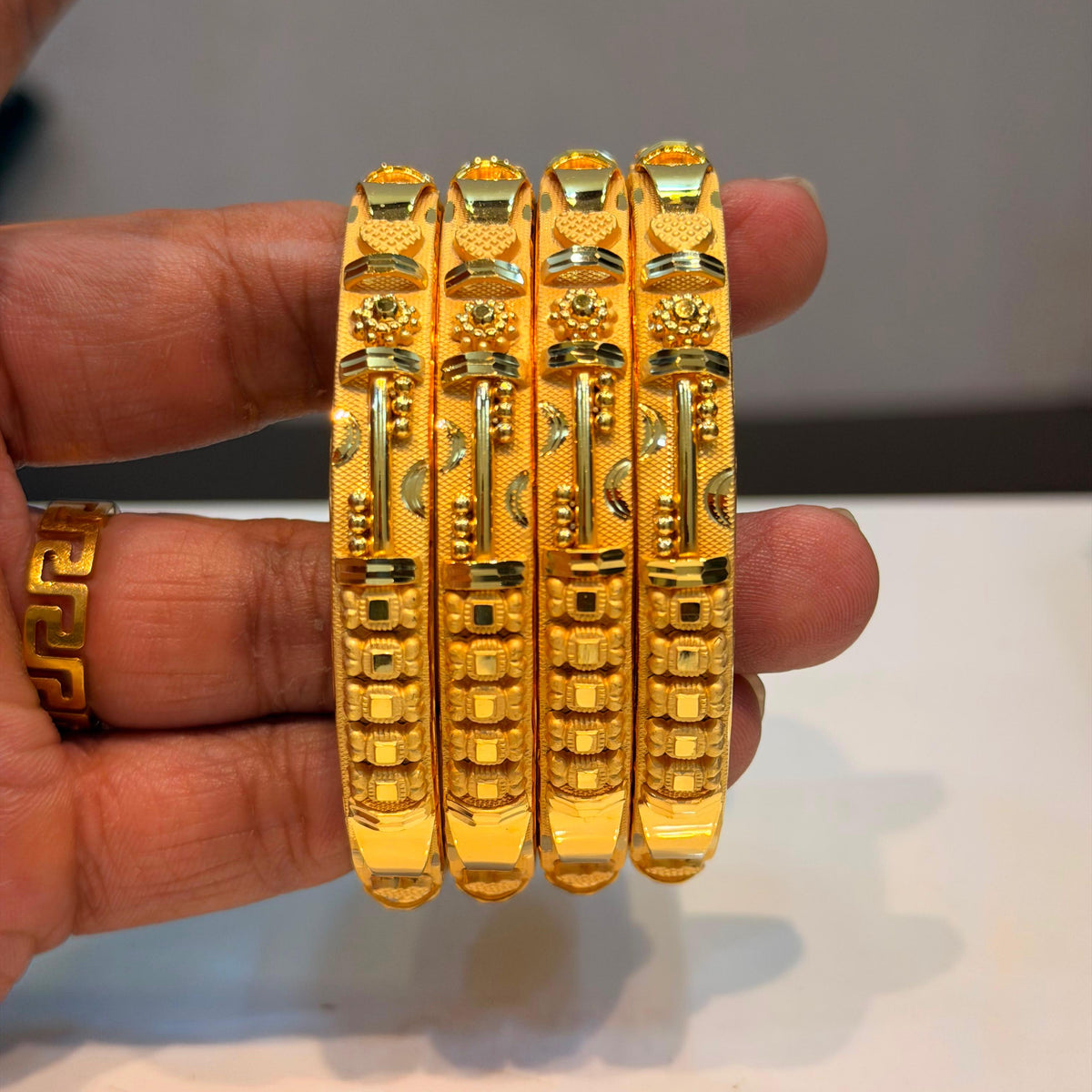 Four ornate gold bangles with detailed geometric and floral engravings, held in a person's hand against a neutral gray background.