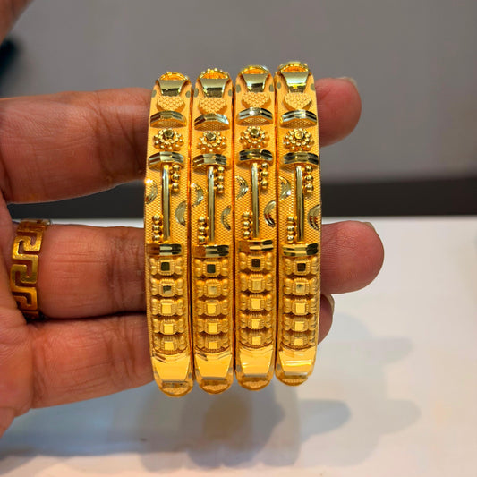 Four ornate gold bangles with detailed geometric and floral engravings, held in a person's hand against a neutral gray background.