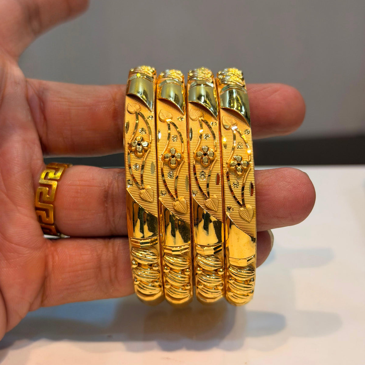 Four ornate gold bangles with detailed floral engravings and small clear stones, held in a person's hand against a neutral background.