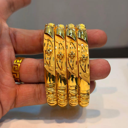 Four ornate gold bangles with detailed floral engravings and small clear stones, held in a person's hand against a neutral background.