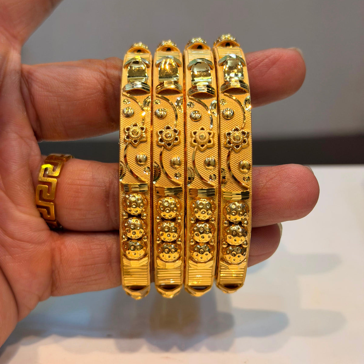 Four ornate gold bangles with intricate floral and geometric patterns, held in a hand. A matching gold ring is also visible on the ring finger.
