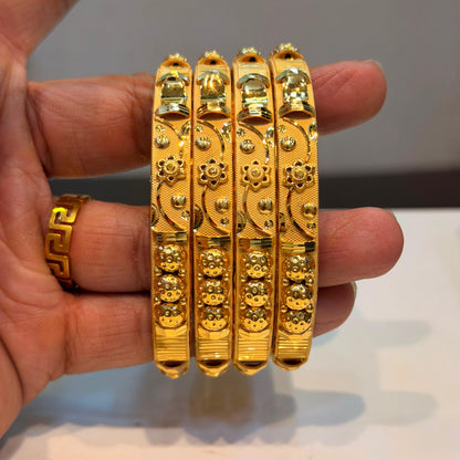 Four ornate gold bangles with intricate floral and geometric patterns, held in a hand. A matching gold ring is also visible on the ring finger.